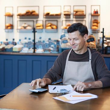 Baker calculating finances at a table in a bakery with bread in the background.
