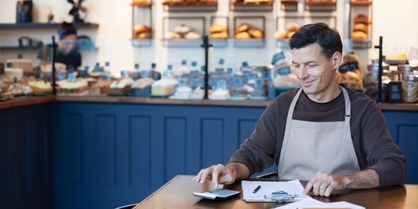 Baker calculating finances at a table in a bakery with bread in the background.