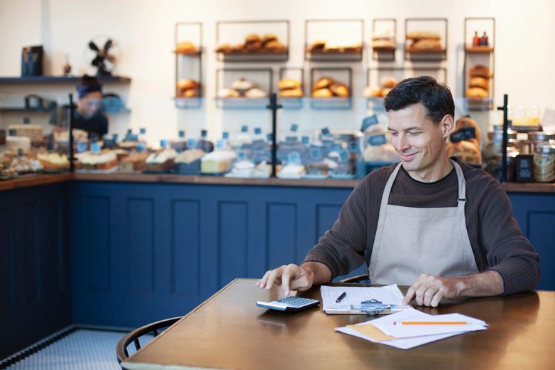 Mature man owner staff employee using calculator for sales analysis accounting bookkeeping in small business bakery cafe