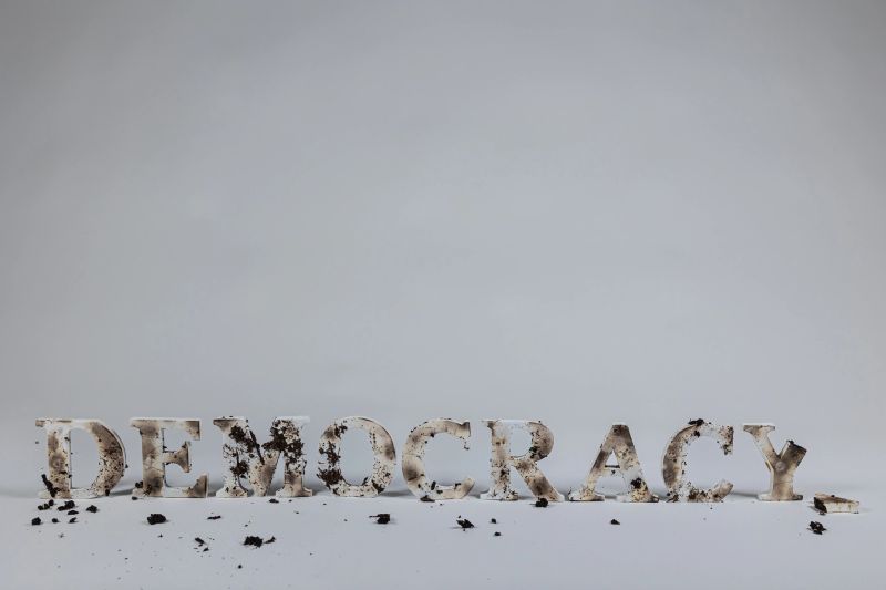 Wood letters spell out the word democracy against a white seamless background.  The letters are burned and muddied, indicating that democracy is under attack.