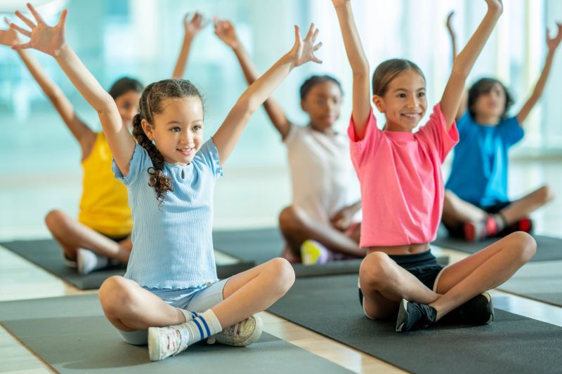 A small group of school aged children sit spread apart on yoga mats as they participate in a mindfulness class together.  They are each dressed comfortably and are sitting cross-legged  with their hands in the air.
