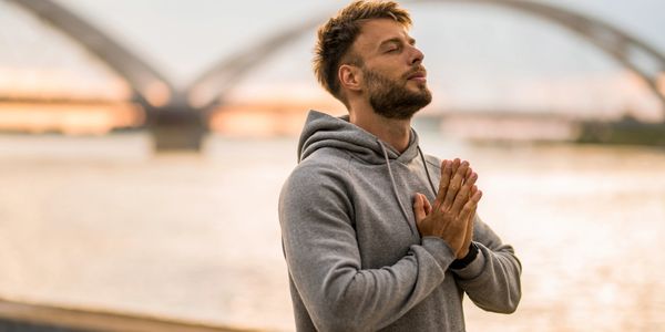 Man in gray hoodie meditates near water with bridge in background.