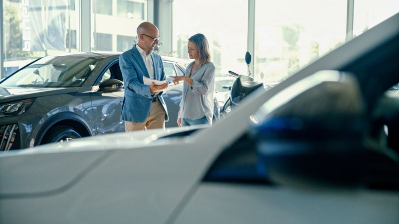 Close-Up of Brand New White Car with Female Customer and Male Salesperson Discussing Documents in the Background at Modern Showroom