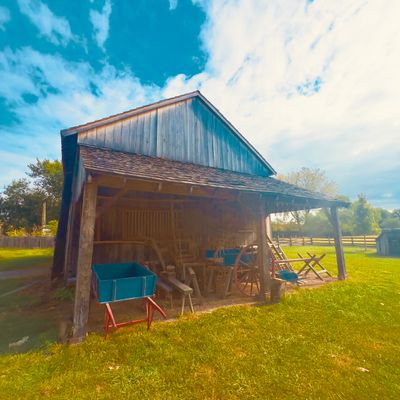 Rustic wooden barn with wheelbarrows and farming tools under a porch on a sunny day.
