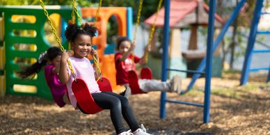 Three children happily swinging on red swings at a playground on a sunny day.