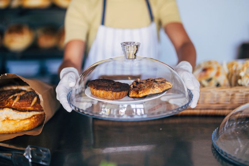 A baker wearing gloves displays two delicious pastries under a glass dome, showcasing freshness in a cozy bakery setting.