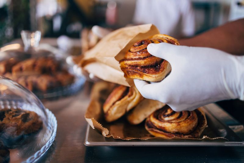 A close-up of a freshly baked cinnamon roll being served by a gloved hand in a bakery. The image captures the warmth and inviting atmosphere of a pastry shop.