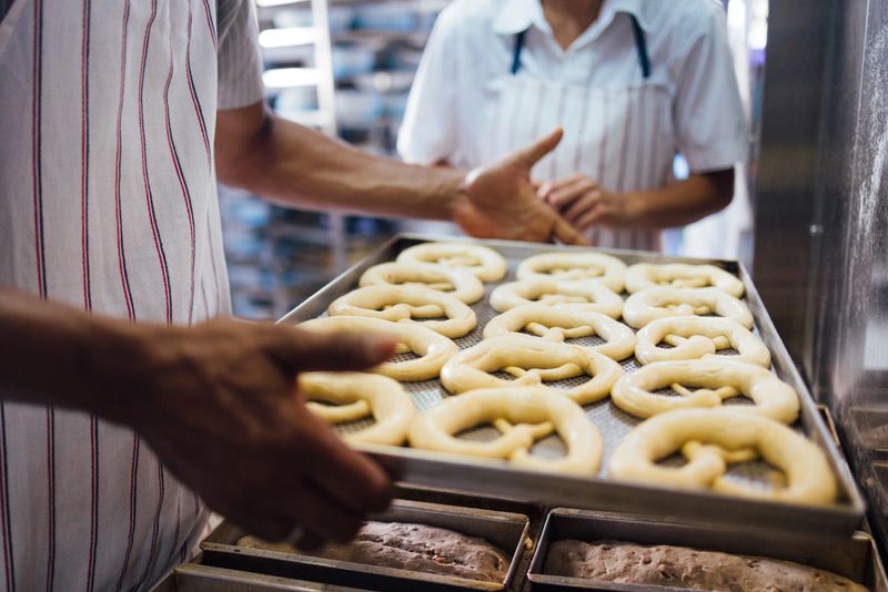 Close-up image of bakers handling a tray of freshly prepared pretzels, showcasing craftsmanship and teamwork in a busy bakery setting.