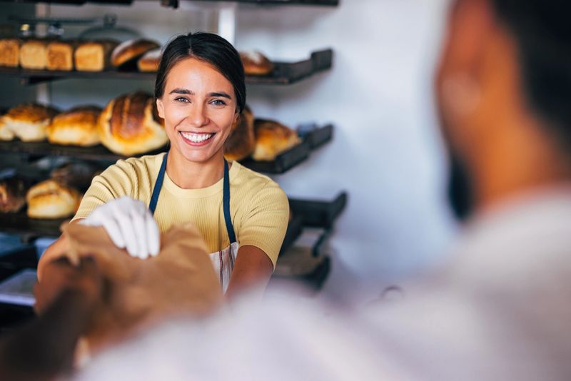 A cheerful baker in an apron hands a paper bag to a customer inside a cozy bakery filled with freshly baked bread.