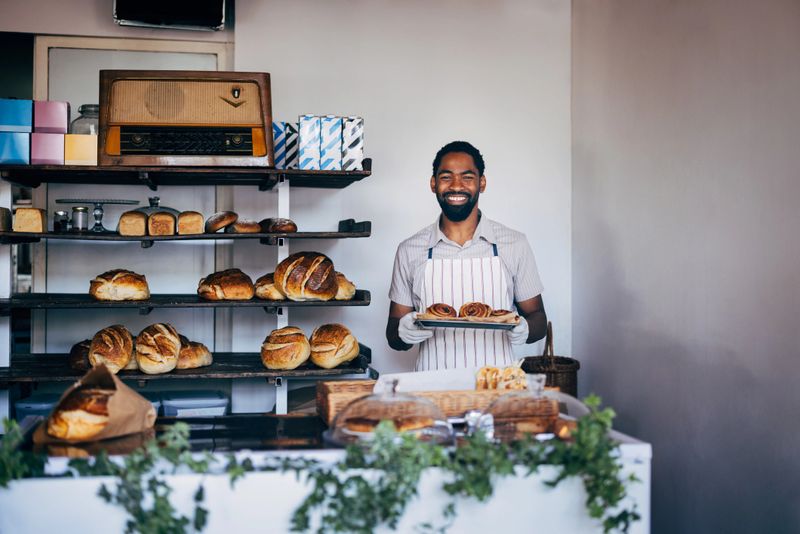 A smiling baker proudly displays fresh pastries in a cozy, rustic bakery. The shelves behind are lined with assorted bread loaves, creating a warm and inviting atmosphere.