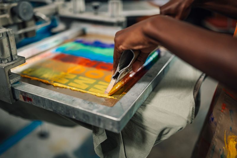 Close up of multicultural female printing workshop worker's hands pressing ink into textile product with squeegee at factory. Cropped picture of unrecognizable printing shop worker printing on t-shirt