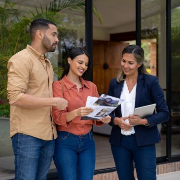 Pareja revisando folletos con agente inmobiliaria frente a casa.