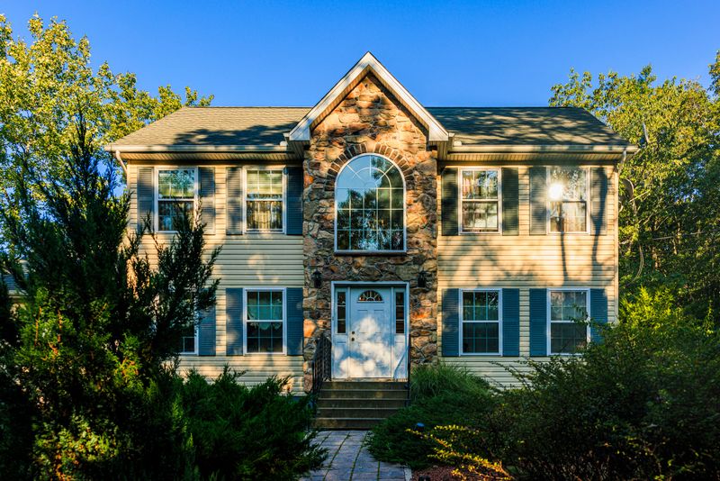 Two-story suburban house with distinctive stone facade and arched window. Beige siding, blue shutters, and green roof harmonize with natural surroundings. Lush greenery frames property, mature trees and shrubs add privacy. Stone steps lead to white front door, creating inviting entrance. Well-maintained landscaping and clear blue sky evoke peaceful, idyllic neighborhood in Poconos region of Pennsylvania.
