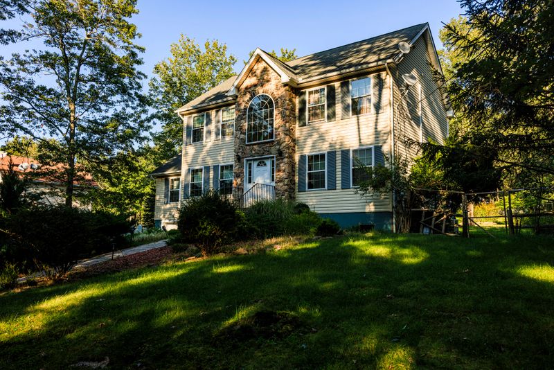 Two-story suburban house with beige siding and white trim stands amid lush green surroundings. Home features prominent arched window above entrance and multiple regular windows. Neatly maintained lawn stretches in front, dotted with trees and shrubs. Property exudes tranquility and comfort, typical of middle-class neighborhoods in Pocono Mountains region of Pennsylvania.