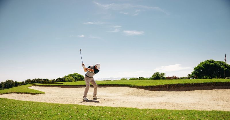A male golfer skillfully swinging a golf club to escape a bunker during a competitive match, capturing determination and precision.