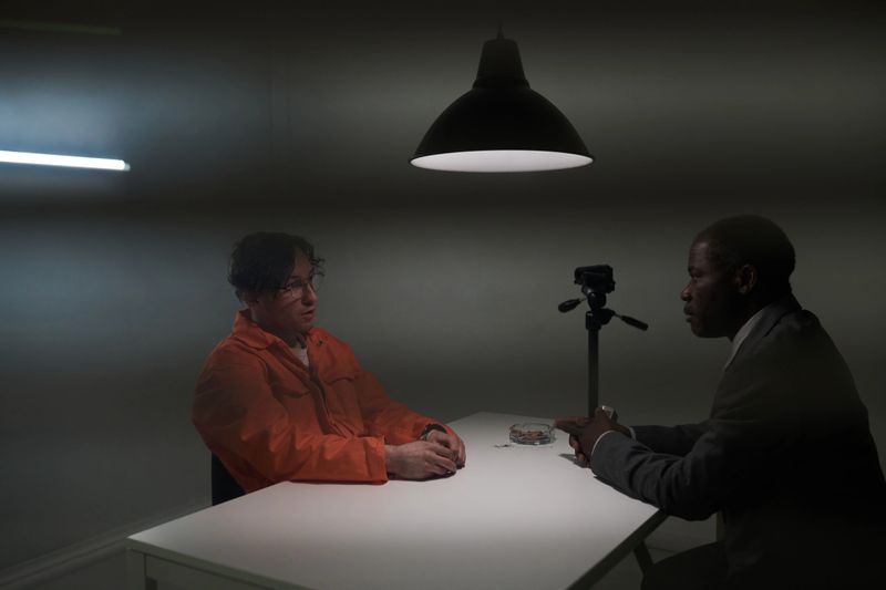 Man wearing orange jumpsuit sitting at table during interrogation session facing another man in suit both men deep in conversation under bright light