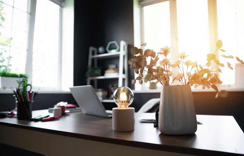 Bright, modern workspace with a laptop, desk accessories, a glowing light bulb, and a vase of flowers. Sunlight pours through large windows, casting a warm, inviting glow over the minimalist setting