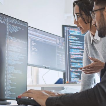 Two programmers collaborate while reviewing code on multiple monitors in an office.