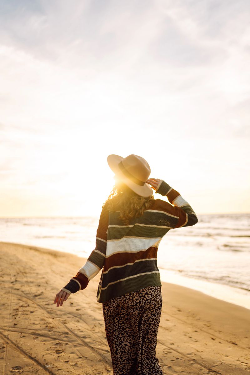 Young woman enjoying a sunset at the beach while wearing a cozy striped sweater, smiling as waves gently lap at the shore. Travel, weekend, relax and lifestyle concept.