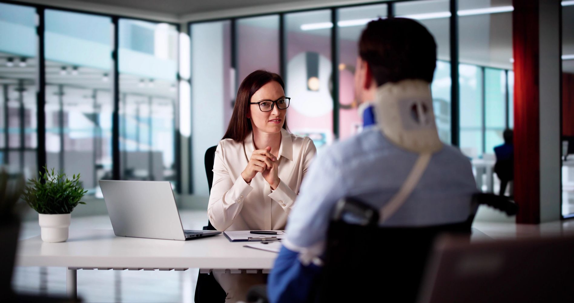 A woman in glasses talks to a man in a neck brace in an office.