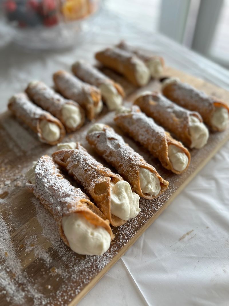 A delectable display of freshly made cannoli, generously filled with a rich and creamy filling, dusted with powdered sugar. Served on a rustic wooden tray, this image captures the allure of homemade Italian desserts in a light-filled setting, perfect for a sweet treat during any gathering.