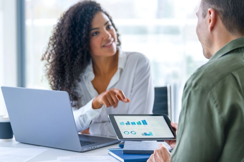 Business man and business woman in a meeting at the office. There is a laptop on the table and the man is holding a digital tablet with finance chart and graphs. The woman happy and smiling and listening