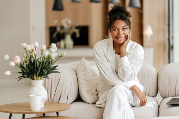 Smiling woman in white sitting on a beige couch with flowers on the table.