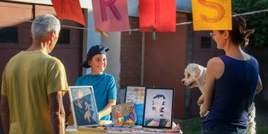 Niña muestra arte en un puesto al aire libre en una feria para dos adultos con perro.