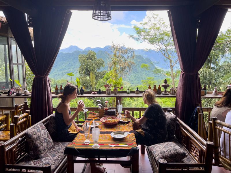 Two female tourists are seated at a table eating a meal. The table has traditional women textile cloth, in colourful warm tones of orange and yellow. Our main characters look out onto a lush green  mountainous valley, the epitome of luxury. The guests  sit on handmade bamboo furniture with woven heritage cushions. This is a bespoke , immersive vacation featuring simplicity and immersion in beautiful nature. A bespoke vacation.