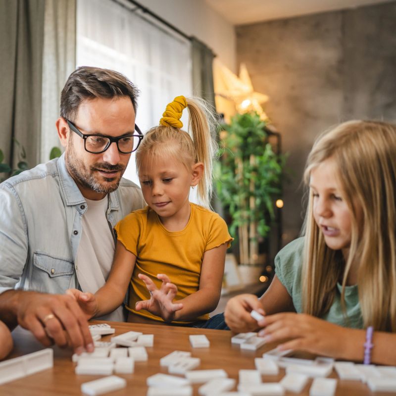 A smiling single father enjoy in a fun and educational game of dominoes with his three young daughters at a wooden table, capturing a joyful family bonding moment at home