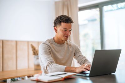 Young man working on a laptop at a table with a notebook.