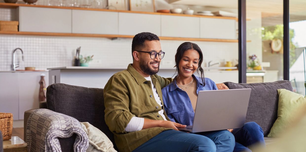 Couple sitting on a couch smiling while using a laptop in a cozy living room.