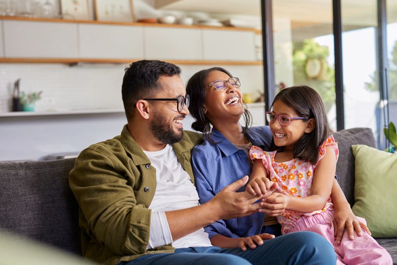 Cheerful middle eastern family with eyeglasses of three having fun together at home. Mixed race parents talking with their little girl in living room. Cheerful family with specs embracing and playing with beautiful indian daughter girl in arms.