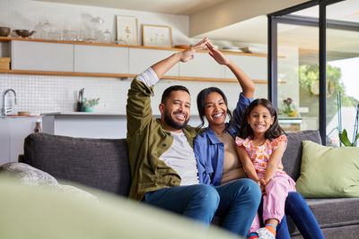 Happy family sitting on a couch, smiling and making a roof shape with their hands in a cozy living room.