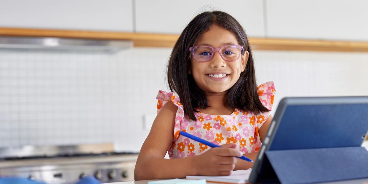 Smiling girl with glasses using a tablet and writing in a notebook.