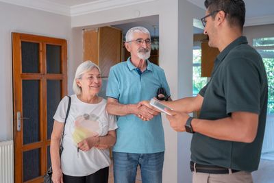 An elderly couple shakes hands with a man holding a tablet in a home setting.