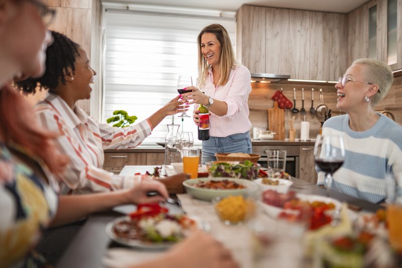 A lively dinner party unfolds as a smiling host serves colorful vegan dishes to her delighted friends, all enjoying a warm evening together in a stylish kitchen.