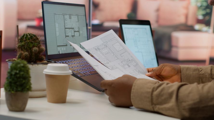 Person reviewing architectural floor plans with laptops and plants on desk.