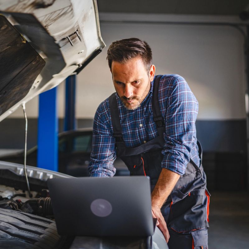 Adult man car mechanic stand by a car with the hood open, use a laptop for diagnostics in a modern workshop garage, highlighting the integration of technology in automotive repairs