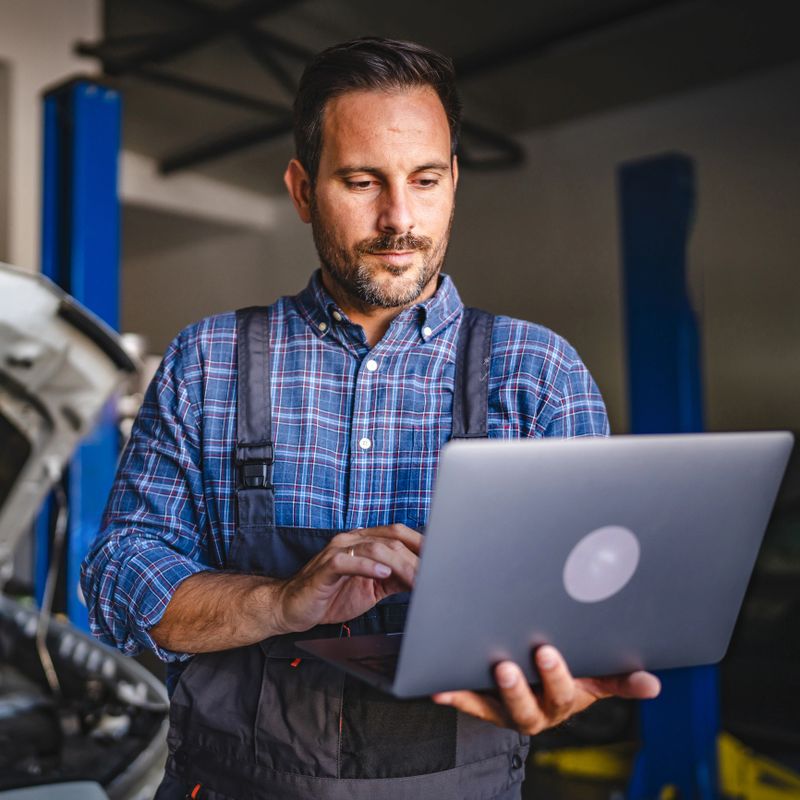 A focused adult man car mechanic in a plaid shirt operates a laptop beside an open-hood vehicle in a modern garage, suggesting the blend of technology and automotive repair