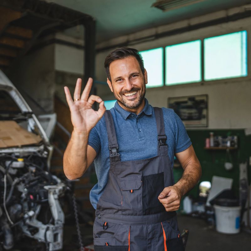 A cheerful car mechanic in a blue polo and overalls gives an okay sign while standing next to a car in a workshop, illustrating positive and professional service