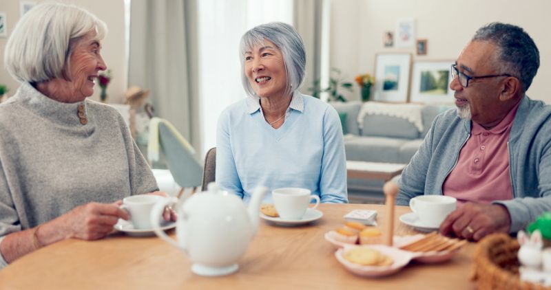 Talking, tea and senior group at table for conversation, snacks and retirement together in house. Happy friends, party and man speaking with elderly women for breakfast, communication and bonding
