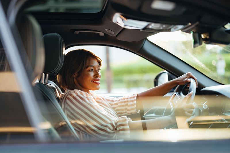 A joyful Black businesswoman confidently driving her car through the city streets. The sunlight highlights her focused expression and professional attire, embodying independence and a sense of freedom.