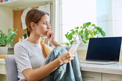 Teen girl seeking mental health support sitting beside a computer and holding a phone.