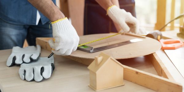 Two people measuring and marking a round wooden board with tools and gloves on a table.