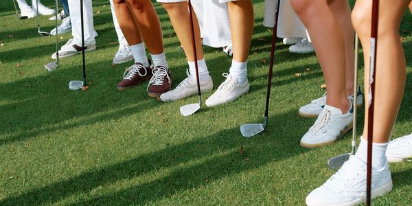 Group of people standing on grass holding golf clubs, wearing casual shoes and socks.