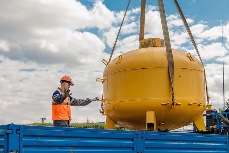 A construction worker in safety gear securing a large yellow industrial tank with a crane, demonstrating industrial safety protocols on a clear and sunny day.
