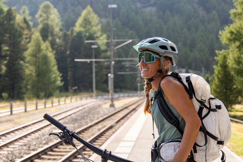 Smiling woman cyclist with mountain bike and backpack at a remote train station surrounded by lush greenery and forested mountains.