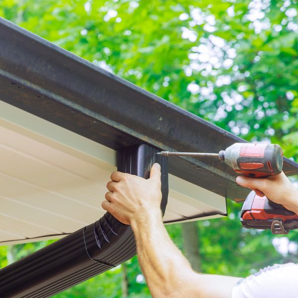 Person installing a black rain gutter on a house roof with a power drill.