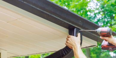 Person installing a black rain gutter with a power drill on a house.